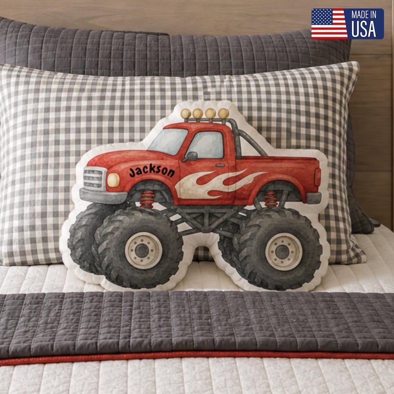 May include: A shaped throw pillow featuring a red monster truck with large black tires, a roll bar with four yellow lights, and white flame graphics on the side. The name Jackson is printed in black script on the side of the truck cab.