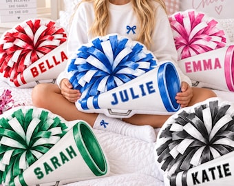 May include: Five plush pillows shaped like cheerleading megaphones with attached pom-poms. Each pillow features a unique color scheme and a name printed in bold block letters: Bella in red, Julie in blue, Emma in pink, Sarah in green, and Katie in black.