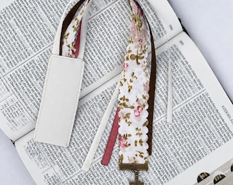May include: A multi-layered bookmark featuring a white faux leather tab, a white fabric strip with a pink and green floral pattern and scalloped edges, a solid brown ribbon, and a thin maroon ribbon, finished with a bronze-toned metal cross charm.