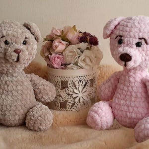 May include: Two crocheted teddy bears, one in beige and one in pink, sitting on a light brown fuzzy surface. The bears have black eyes and brown noses.