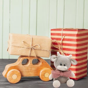 May include: A gray crocheted cow toy with a pink bow tie sits in front of two wrapped gifts. One gift is brown with twine and the other is red and white striped. The gifts are sitting on a wooden surface.