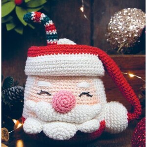 Puede incluir: Una funda de taza de ganchillo de Papá Noel. La funda es blanca con un gorro y ribete rojos, una nariz rosa y una barba blanca. El asa es roja. El fondo incluye decoraciones navideñas.