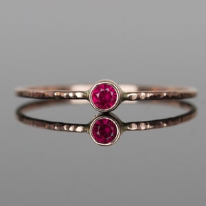 May include: A delicate ring with a hammered band and a bezel-set, round, ruby-red gemstone. The ring is displayed on a reflective surface, showcasing its design. The band has a textured, hammered finish.