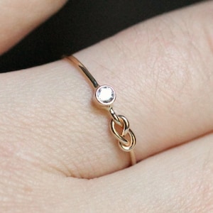 Könnte beinhalten: Ein zarter Goldring mit einem kleinen, klaren Stein und einem Knotenband.