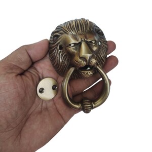 May include: Brass lion head door knocker with a ring handle. The knocker is in a hand, and a separate round brass plate with two holes is visible.