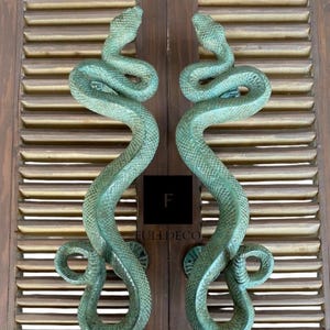 Pu&ograve; includere: Un paio di maniglie per porte a forma di serpente verde antico. Le maniglie sono in metallo e hanno un design di serpente realistico. Le maniglie sono montate su una porta in legno.