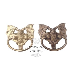 May include: Two brass bat door knockers, one with a polished finish and the other with a more rustic finish. The knockers are shaped like bats with outstretched wings and have a ring pull.