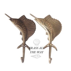 Pode incluir: Dois ganchos de parede de metal dourado em forma de peixe-espada. Os ganchos são em forma de peixe-espada com as barbatanas abertas. O texto "BRASS ALL THE WAY" está impresso abaixo dos ganchos.