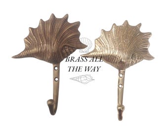 Può includere: Due ganci in ottone a forma di conchiglia con finitura testurizzata. I ganci sono montati su uno sfondo bianco. "BRASS ALL THE WAY" è stampato in nero sotto i ganci.
