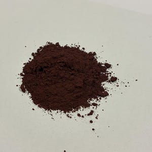 May include: A small, irregular pile of fine, dark reddish-brown powder. The texture appears granular and dry, with scattered loose particles extending from the main cluster, creating a natural, uneven shape on the flat, neutral-colored surface.