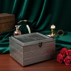 May include: A silver-colored jewelry box with an ornate, embossed design on the lid and sides. The box has a clasp closure and sits on a wooden surface. Red roses are in the foreground, with a green velvet backdrop.