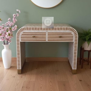 Könnte beinhalten: Ein weiß-braun gestreifter Schreibtisch mit zwei Schubladen und goldfarbener Hardware. Der Schreibtisch hat abgerundete Kanten und goldene Füße. Eine weiße Vase mit rosa Blumen und eine kleine Pflanze auf einem Holzständer stehen neben dem Schreibtisch.