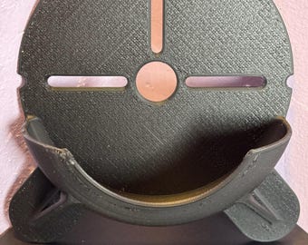 May include: A black, semi-circular object with a textured surface. It features a curved lower section and a central hole with a vertical slot above it. Two horizontal slots are on either side of the central hole. The object is mounted on a black surface.