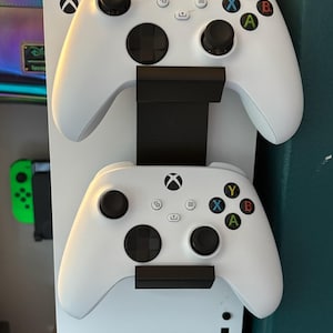 May include: A white Xbox controller stand holding two white Xbox controllers. The stand is black and has the Xbox logo. The controllers have black buttons and colored action buttons.