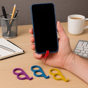 May include: A blue smartphone with a red finger grip is held in a hand. Three coloured finger grips in purple, blue, and yellow are on a wooden desk. A white mug of coffee, a notebook, and office supplies are also visible.
