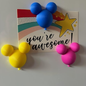 May include: Three Mickey Mouse-shaped balloon magnets in yellow, pink, and blue. A rectangular sign features a rainbow, star, and the words "you're awesome!". The magnets are attached to a white surface.