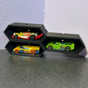 Puede incluir: Tres estantes hexagonales negros que exhiben coches de juguete. Los coches son amarillos, verdes y amarillos con detalles en rojo. Los estantes están dispuestos en una esquina, con los coches visibles desde el frente.