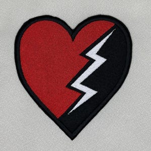 Pode incluir: Um coração vermelho com um raio preto e branco que o divide ao meio. O coração é bordado em um tecido branco.