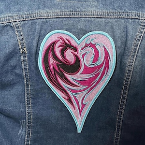 May include: A heart-shaped patch featuring two stylized dragons facing each other. The dragons are black and magenta, outlined in light blue, and set against a pink background. The patch is sewn onto a denim jacket.