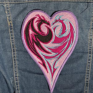 May include: A heart-shaped embroidered patch featuring two dragons facing each other. The dragons are black and magenta with teal accents, set against a light purple background. The patch is sewn onto a denim jacket.