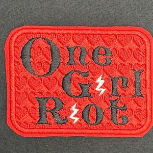 May include: A rectangular red embroidered patch with the words "One Girl Riot" in black, with lightning bolt graphics. The background of the patch is a repeating heart pattern. The patch is likely for clothing or accessories.