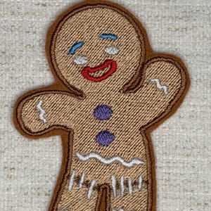 May include: A brown gingerbread man patch with a stitched design. The patch features a smiling face with blue tears, a red mouth, and purple buttons. White zigzag lines and vertical lines add detail to the design.