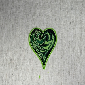 May include: A heart-shaped patch with a green dragon design. The patch has a black interior with a green dragon silhouette and a bright green border. The patch is on a light gray fabric background.