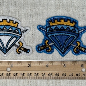 May include: Two embroidered patches featuring a diamond shape with a crown and crossed swords. The patches are blue, white, and gold. A ruler shows the patches are approximately 2 inches wide.