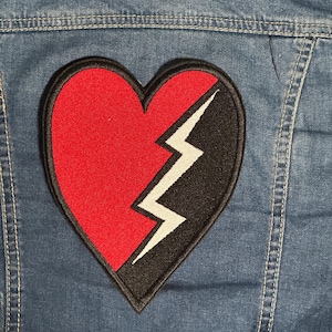May include: A denim jacket with a large embroidered patch of a broken heart. The heart is divided into red and black sections, with a white lightning bolt design. The patch is outlined in black.