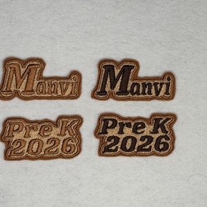 May include: Four embroidered patches featuring the text Manvi and Pre K 2026 in two different color variations. The patches have a tan background with brown stitching and a dark brown border, showcasing clear lettering and a textured fabric finish.