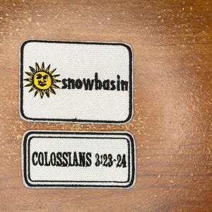 May include: Two rectangular patches on a wooden surface. The top patch is white with a black border, featuring a yellow sun graphic and the word "snowbasin". The bottom patch has the text "COLOSSIANS 3:23-24".