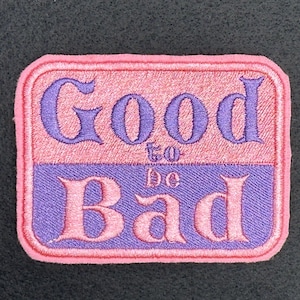 May include: Embroidered patch with the words "Good to be Bad" in purple script on a pink and purple background. The patch has a pink border and is rectangular in shape.