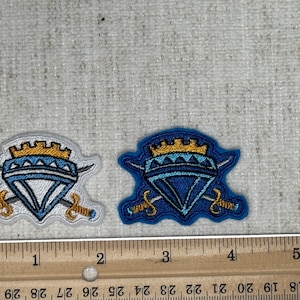 May include: Two embroidered patches with a crest design. Each patch features a blue and gold crown above a diamond shape, crossed swords, and a white or blue background. A ruler shows inches.