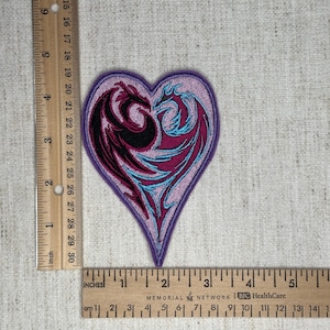 May include: A heart-shaped patch with a purple border. Inside, two stylized dragons intertwine, one black and the other magenta with turquoise accents. The patch measures approximately 3 inches wide.