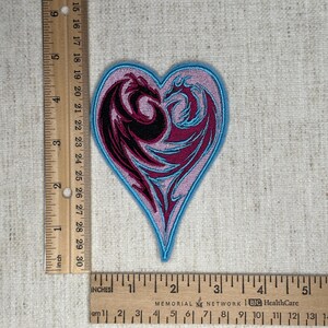 May include: Embroidered heart-shaped patch featuring two dragons facing each other. The dragons are in shades of black and magenta, outlined in light blue. The patch is approximately 3 inches wide and 3 inches tall.