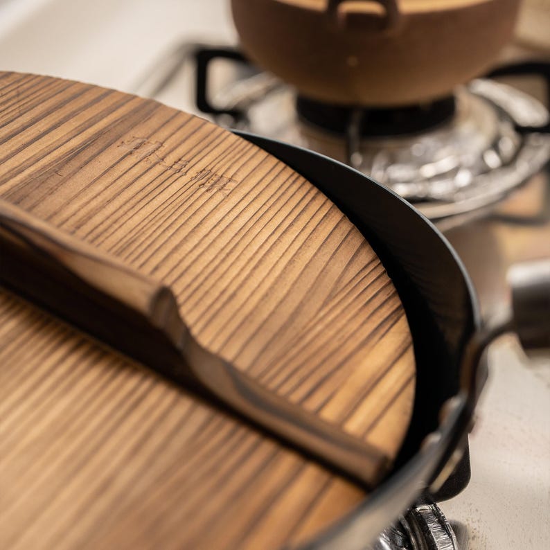 May include: A wooden lid with a handle sits on top of a black cast iron pot. The lid has a natural wood grain and is slightly curved.