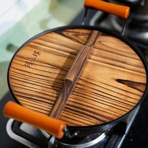 May include: A black wok with a wooden lid. The lid has a wooden handle and is marked with Japanese characters. The wok is on a stovetop.