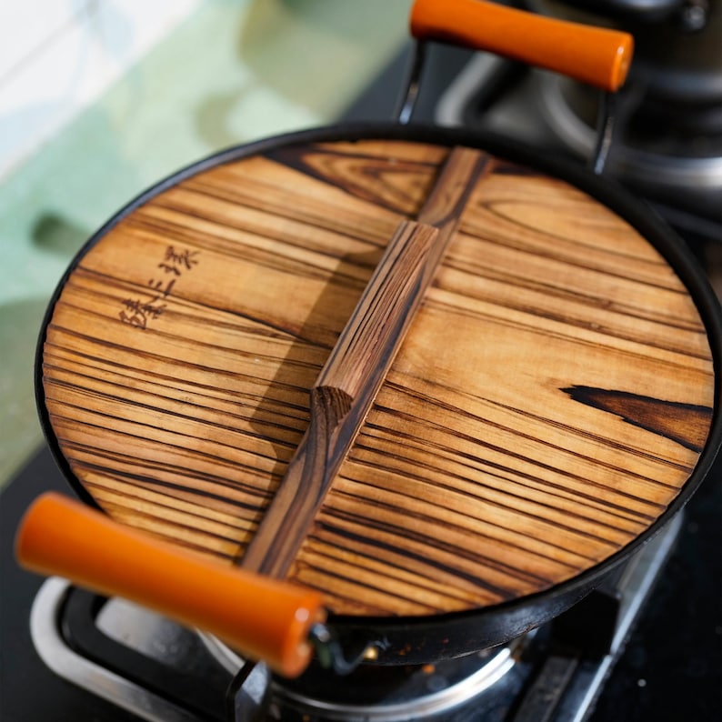 May include: A black wok with a wooden lid. The lid has a wooden handle and is marked with Japanese characters. The wok is on a stovetop.