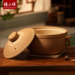 May include: A tan ceramic cooking pot with a lid, set on a wooden surface. The lid is slightly ajar, revealing the pot's interior. The lid features a decorative floral design and a small knob. The pot has two handles.