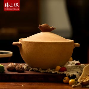 May include: A tan ceramic cooking pot with a lid and two side handles. The lid is topped with a decorative brown bird-shaped handle. The pot sits on a wooden surface, with the text "ZHENSHUANHUAN" in a red box in the upper left corner.