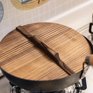 May include: A wooden lid with a dark brown handle sits on top of a black wok. The lid has a wood grain pattern and is slightly domed. The wok is sitting on a stovetop.