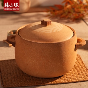 May include: A round, light brown ceramic cooking pot with a matching lid. The lid features a decorative wave pattern and a small, square handle. The pot has two loop handles and sits on a woven mat. The text "ZHENSANHUAN" is visible in the upper left corner.
