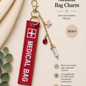 May include: A red fabric keychain tag with white embroidered text reading MEDICAL BAG and a white cross symbol. It features a gold-tone lobster clasp and a gold chain with three charms: a pink bandage, a red teardrop gem, and a white syringe.