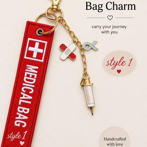 May include: A red fabric keychain with white embroidery reading MEDICAL BAG and style 1, featuring a gold-tone lobster clasp and chain. Attached charms include a white and red bandage, a light blue awareness ribbon, and a white syringe charm.