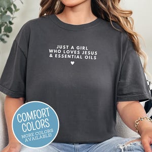 May include: Dark grey t-shirt with the text "JUST A GIRL WHO LOVES JESUS & ESSENTIAL OILS" in white. A small white heart is below the text. A blue circle with the words "COMFORT COLORS" and "MORE COLORS AVAILABLE!" is in the lower left corner.