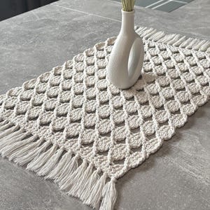Puede incluir: Camino de mesa de macramé color crema con un patrón geométrico y flecos. Un jarrón blanco con un asa única contiene espigas de trigo secas. La mesa es gris.