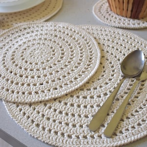 May include: Cream-colored, crocheted placemats and coasters. The placemats are round with a spiral design, and a spoon and fork are placed on top. A white bowl, a napkin holder, and a basket of bread are in the background.