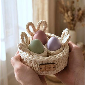 Puede incluir: Una cesta de ganchillo hecha a mano, de color blanco roto, con tres huevos de colores pastel. La cesta tiene detalles de orejas de conejo y una pequeña etiqueta de madera que dice "knit". Una encantadora decoración de Pascua.