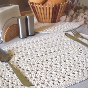 May include: Cream-colored, crocheted placemat with a textured, circular design. A stainless steel spoon rests on the placemat. A basket of bread rolls and a salt and pepper shaker are in the background. The placemat is approximately 12 inches in diameter.