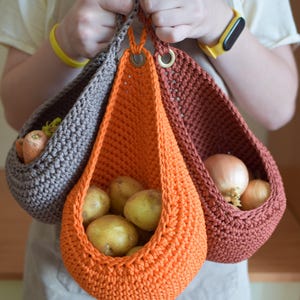 May include: Three teardrop-shaped hanging baskets crocheted from thick cord in shades of gray, bright orange, and terracotta. Each basket features a metal grommet at the top for suspension and contains fresh produce including carrots, potatoes, and onions.
